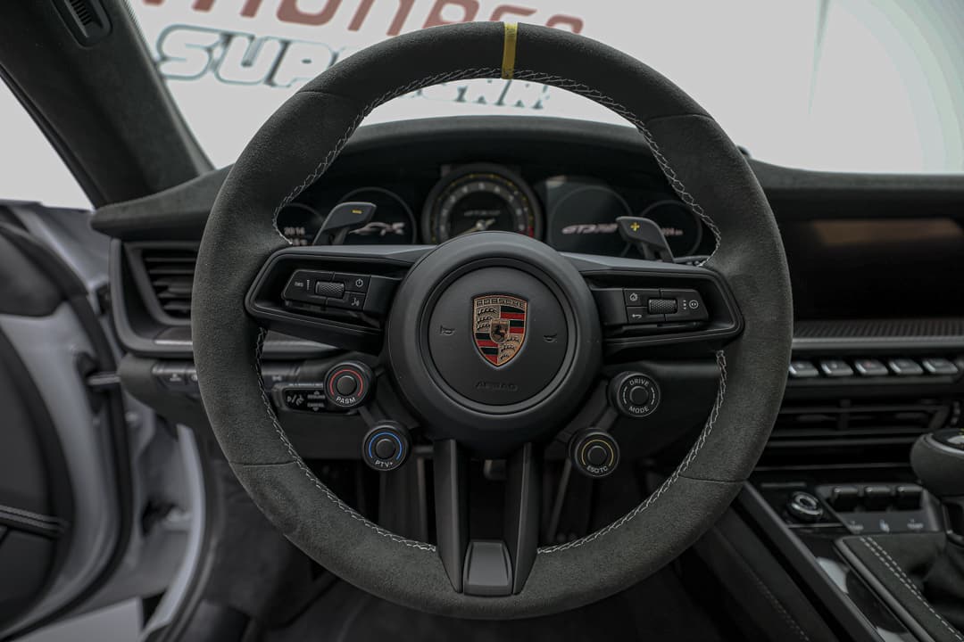 Cette image du volant de la Porsche 911 GT3 RS révèle un design sportif et fonctionnel, avec des commandes intégrées pour un accès facile aux fonctionnalités, une finition en Alcantara pour une prise en main optimale, et un emblème distinctif qui souligne la performance de ce modèle emblématique.