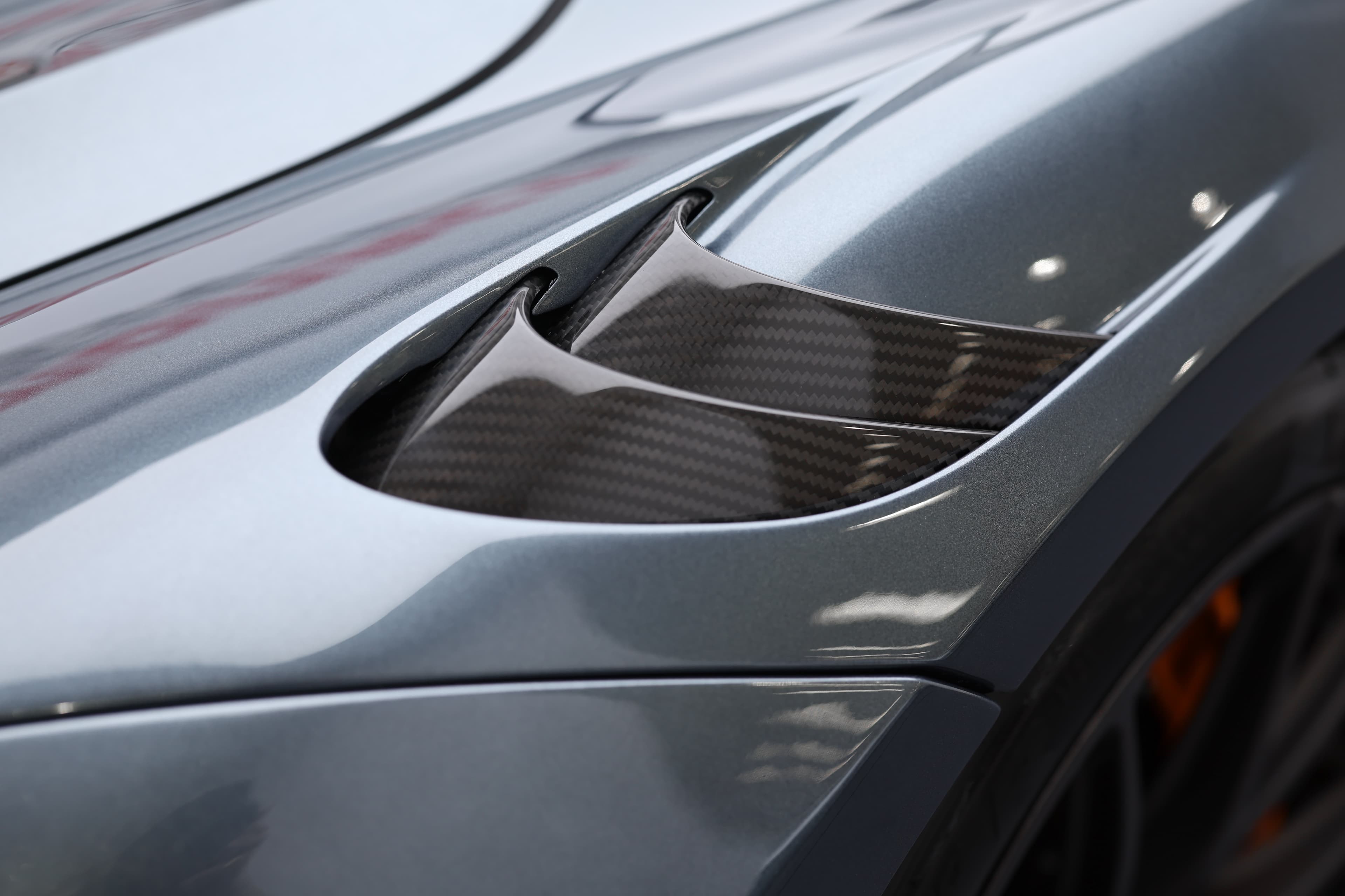 L'image se concentre sur l'entrée d'air en carbone de la McLaren 765LT, mettant en avant le design complexe et fonctionnel qui améliore la circulation de l'air et le refroidissement, accentuant ainsi l'aspect technique et sportif de la voiture.