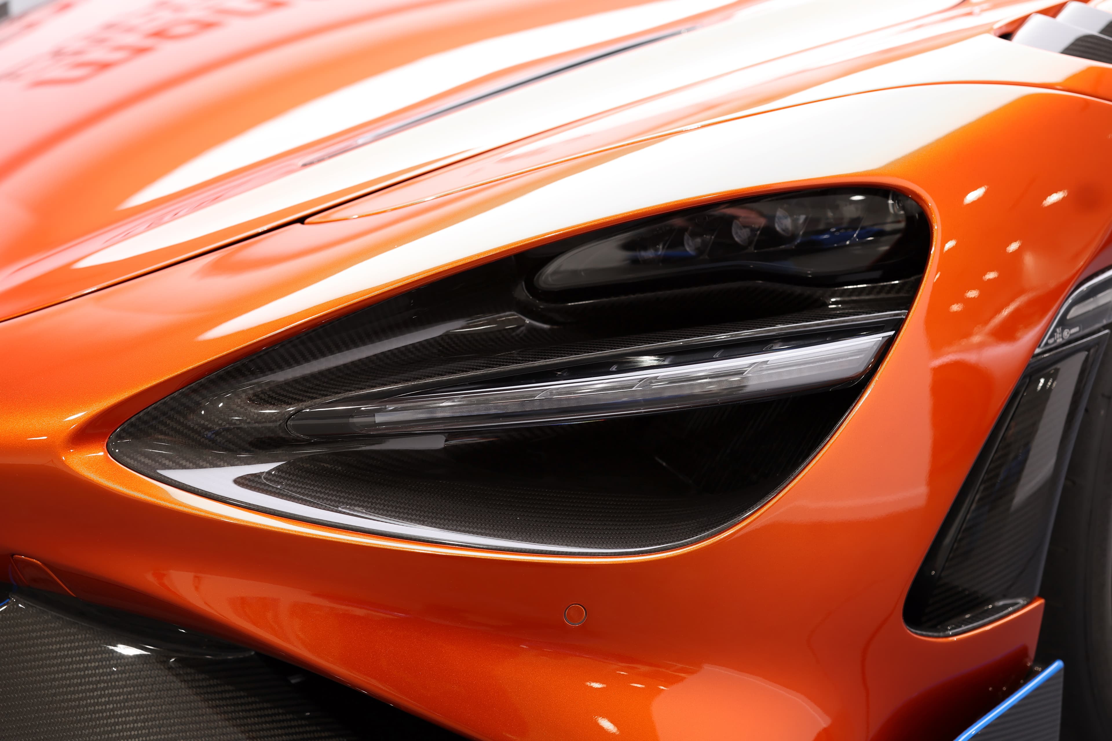 Gros plan sur le phare de la McLaren 765LT, mettant en avant le design dynamique et l'intégration de matériaux en carbone, illustrant l'innovation et l'esthétique moderne de cette supercar.