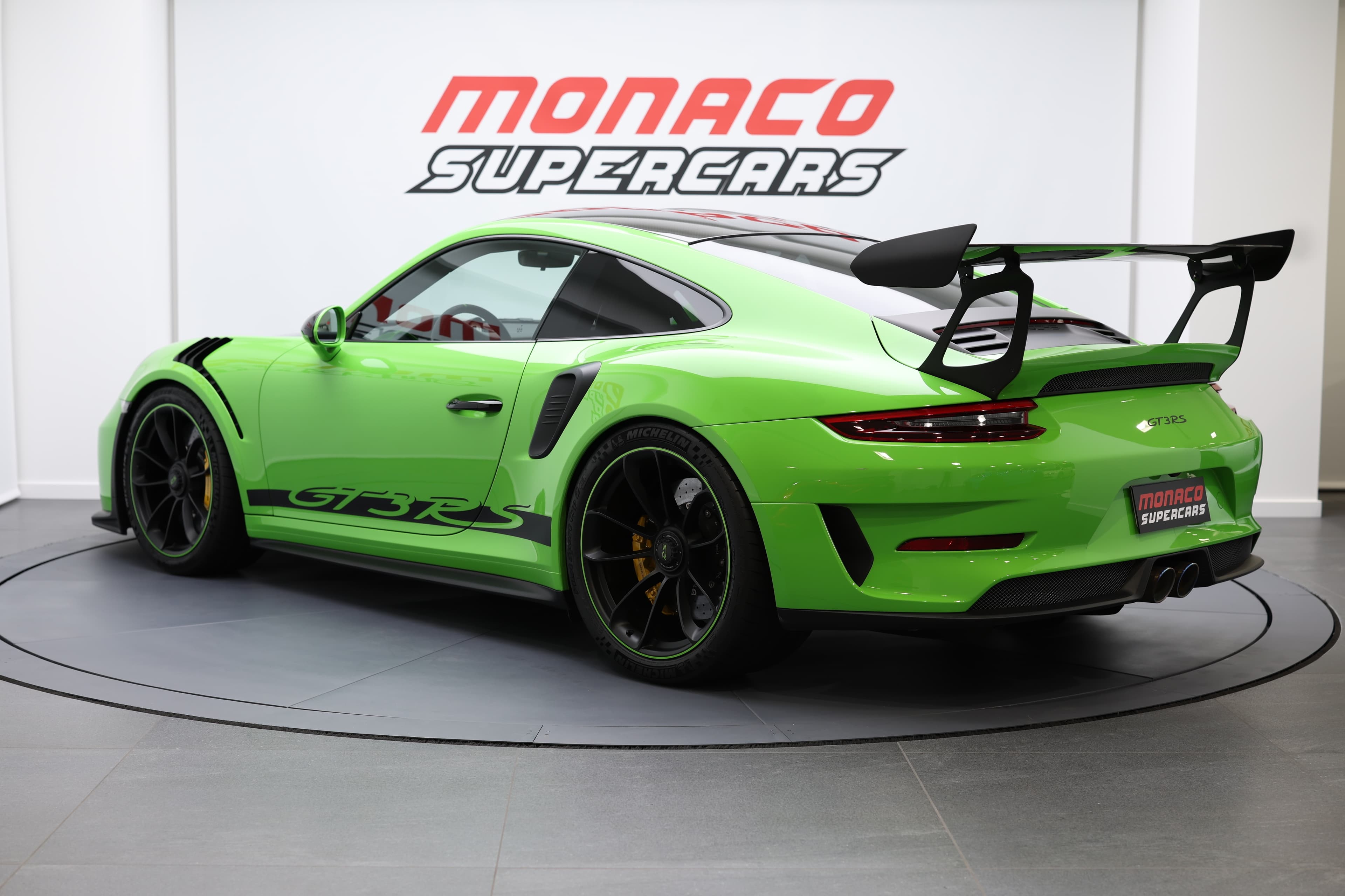 Vue arrière trois-quarts d’une Porsche GT3RS verte exposée chez Monaco Supercars, mettant en valeur son aileron imposant et son design extrême.